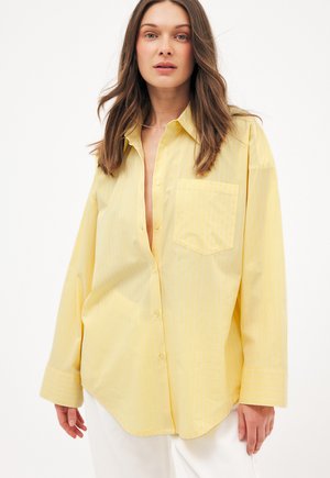 Kobieta o brązowych włosach do ramion, ubrana w luźną, jasnżółtą koszulę zapinaną na guziki i białe spodnie, stojąca na białym tle.