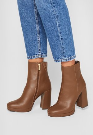 Brązowe skórzane botki na słupku, z bocznym zamkiem i gładką, wypolerowaną fakturą. Noszone do skróconych niebieskich dżinsów.