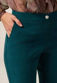 Pantalones de vestir en color azul verdoso, confeccionados con una tela texturizada que presenta un frente plano, bolsillos laterales y un detalle de botón plateado en la cintura.