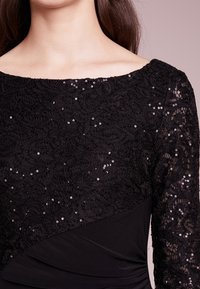 Černé šaty s krajkovou korzetovou částí s vzorem a flitry, dlouhými rukávy a přiléhavou, nařasenou spodní částí. Hladká textura a strukturovaný design.