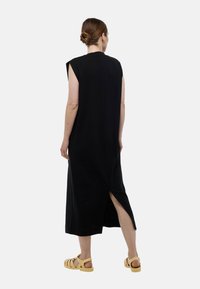 Robe midi noire sans manches avec un décolleté haut au dos et des fentes latérales, fabriquée en tissu doux. Associée à des sandales jaune clair.