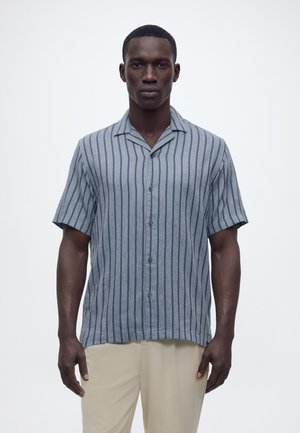 Bărbat de culoare cu păr scurt, purtând o cămașă cu mâneci scurte, albastru-gri, cu dungi verticale negre și pantaloni bej, stând în picioare pe un fundal alb.