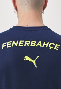 Camiseta de algodón azul marino con la palabra "Fenerbahçe" en letras amarillas destacadas y un logo amarillo de Puma, ubicados en la parte posterior.