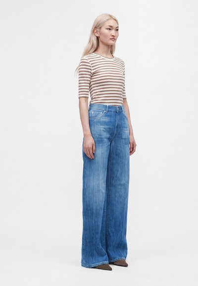 Jeune femme aux longs cheveux blonds porte une chemise rayée beige et blanche et un jean bleu à jambes larges, debout sur un fond blanc uni.