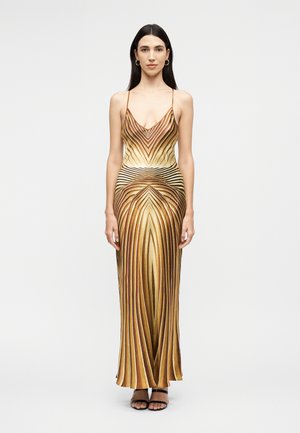 Γυναίκα όρθια, φοράει ένα αμάνικο, μακρύ μέχρι το πάτωμα φόρεμα με χρυσές και καφέ ρίγες, λεπτές τιράντες και μαύρες ανοιχτές γόβες.