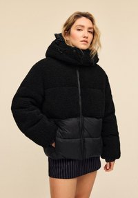 Črna napihnjena jakna z mehkimi teksturiranimi zgornjim delom, gladkim spodnjim delom in velikim kapuco z vrvico za zatezanje.