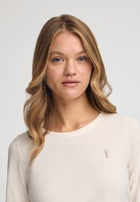 Cremfarbendes Langarmshirt aus weichem Stoff mit einem dezenten, in Gold gestickten Markenlogo auf der linken Brustseite.