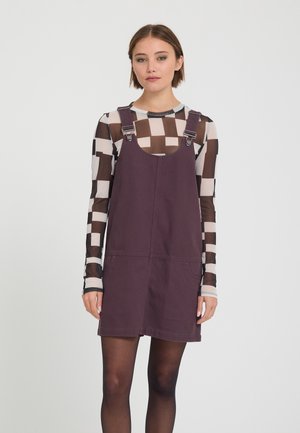 Jeune femme portant une robe sans manches violette par-dessus un top en mesh à manches longues à carreaux noir et blanc, debout devant un fond uni.