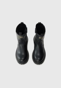 Une paire de bottes noires en cuir montantes avec des semelles épaisses, des zips sur les côtés, et de petites plaques rectangulaires dorées en logo sur la partie supérieure avant.