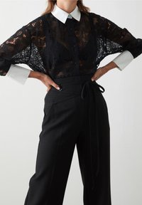 Czarna bluzka z koronki z białym kołnierzykiem, z przodem na guziki i szerokimi rękawami, zestawiona z czarnymi spodniami o wysokim stanie z sznurkiem i teksturowanymi akcentami.