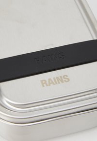 Metalowa pojemnik o szczotkowanej aluminiowej powierzchni, z czarną gumową opaską i wytłoczonym napisem "RAINS" na pokrywie.