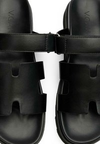 Black vegan leather sandals feature two wide straps with cut-out designs and a textured finish. The sole is smooth with a rounded shape.