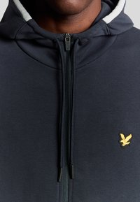 Marineblauwe hoodie met rits, gemaakt van zachte stof, met een wit gevoerde capuchon en een klein geel vogel logo op de borst.