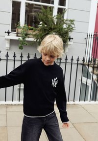 Černý pletený svetr s bílým logem na hrudi, kulatým výstřihem a kontrastním lemem. Kombinován s tmavými džíny. Venku v prostředí zeleně.