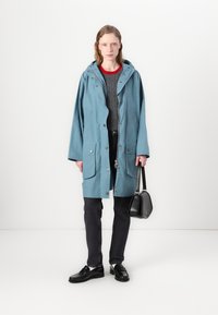 Light blue waterproof parka with a hood, front snap buttons, two front pockets, worn over a gray sweater, paired with dark jeans and black shoes.