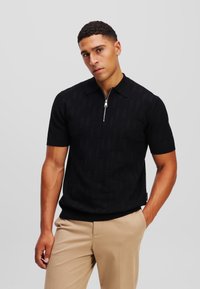 Černé polo tričko s krátkým rukávem a polo zipem, které má texturované geometrické vzory a přiléhavý střih. Tkanina vypadá jako žebrovaná.