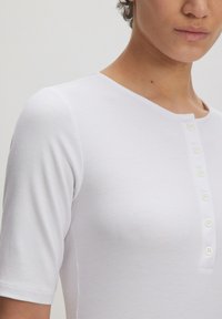 Witte geribbelde top met korte mouwen, voorzien van een ronde halslijn en een knopenlijst met zes witte knopen aan de voorkant.