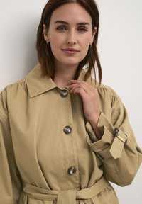 Khaki trenčkot s golierom, gombíkovým zapínaním a opasok. Tkanina je hladká s mierne štruktúrovaným dizajnom.