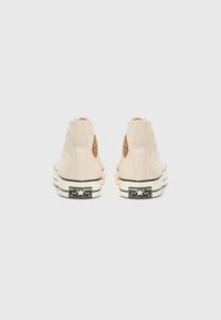 Beige high-top sneakers made of canvas with a textured look, featuring a brown logo patch and black rubber soles.