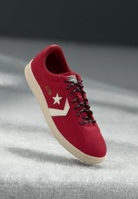 Red suede sneaker with a beige star logo, black laces, and a cream rubber sole. Textured upper and structured design.