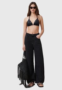 Top bikini triangolare nero, pantaloni larghi neri a vita alta e una borsa intrecciata nera con frange; il modello indossa occhiali da sole neri.