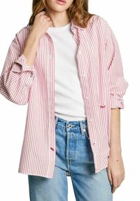 Camicia a righe rosse e bianche con un taglio morbido e maniche arrotolate, abbinata a una maglietta bianca semplice e jeans azzurri chiari.