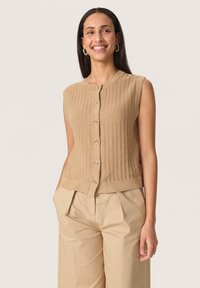 Beige ärmlös cardigan med knäppning fram, texturerat stickat mönster och ribbad nederkant. Matchas med lösa byxor i neutrala färger.