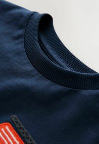 Tumesinine puuvillane dressipluus soonikkoes ümber kaela. Esiküljel on tikandiga muster oranzi ja musta värviga. Pehme ja sile tekstuur.