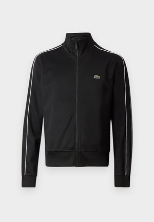 Jachetă sport neagră, cu fermoar, confecționată dintr-un material neted, având dungi albe pe laterale și un mic logo verde pe piept. Design cu guler înalt.