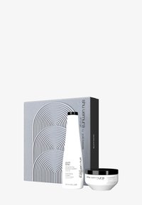 Bouteille de shampooing blanche et pot d'accent noir dans une boîte grise embossée présentant des motifs de lignes courbes. Des étiquettes de texte indiquent les détails du produit.