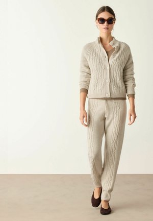 Kvinde iført beige kabelstrikcardigan og matchende bukser med brune flade sko og skildpaddemønstrede solbriller står på en neutral baggrund.