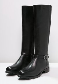 Paire de bottes en cuir noires montantes jusqu'aux genoux, avec des talons bas et des boucles sur le côté, debout verticalement sur une surface blanche contre un fond clair.