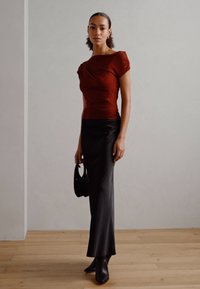 Rustfarget tettsittende topp med drapert design, sammen med et elegant svart satin midi-skjørt. Modellen holder en svart håndveske og har på seg spisse ankelstøvletter.