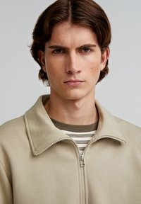 Light beige zip-up jacket with a high collar, smooth texture, and contrasting zipper. Worn over a striped crewneck shirt.