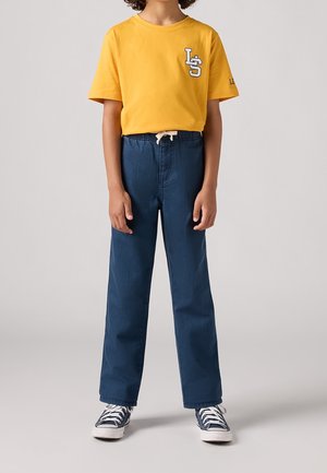 Dziecko o kręconych włosach w żółtej koszulce z krótkim rękawem, niebieskich szerokich spodniach ze ściągaczem oraz granatowych tenisówkach z białymi sznurówkami.