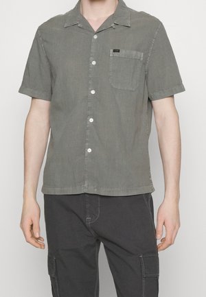 Hombre vestido con una camisa gris de manga corta con botones y bolsillo en el pecho, y pantalones cargo oscuros, de pie con los brazos relajados a los lados.