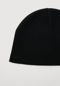 Melnā adīta cepure ar smalku "HUGO" logo izšuvumu tuvu mala uz vienkārša gaiša fona.