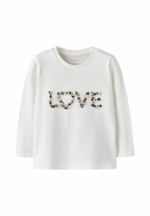 Chemise blanche à manches longues avec le mot "LOVE" sur le devant, constitué de petits détails en tissu noir et beige.