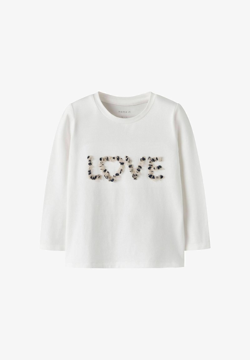 Chemise blanche à manches longues avec le mot "LOVE" sur le devant, constitué de petits détails en tissu noir et beige.