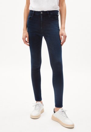 Osoba ubrana w ciemnoniebieskie obcisłe dżinsy i białe trampki z beżowymi podeszwami, stojąca na tle gładkiego tła.