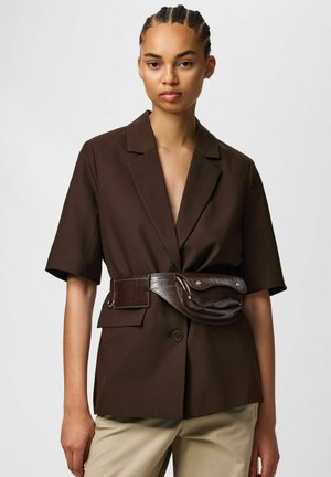 Jonge vrouw draagt een bruin kortermig colbert met een bijpassende bruine heuptas en beige broek tegen een effen lichte achtergrond.