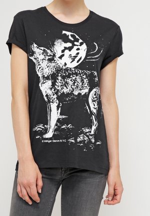 Sort t-shirt med hvid grafik af en ulv, der hyler foran en fuldmåne og klippebund, båret af en person i grå jeans.