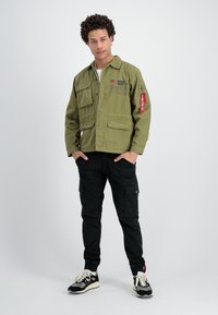 Chaqueta verde oliva con múltiples bolsillos frontales, cierres de botón y un acento en una etiqueta roja. Pantalones cargo negros y zapatillas negras.