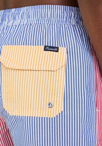 Costumi da bagno a righe con sezioni blu, rosse e gialle, dotati di una tasca gialla, di un occhio in metallo e di un'etichetta con il logo sulla patta della tasca.