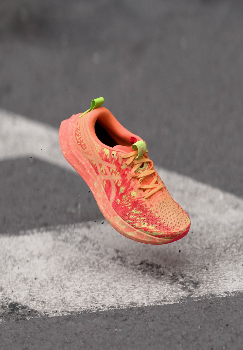 Baskets de course avec un upper en tricot rose vif et orange, des accents verts et une semelle texturée, en suspension au-dessus d'une surface grise.