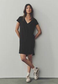Sort, kortærmet kjole med V-udskæring og sideslidser, lavet af et glat materiale. Båret med hvide sokker og beige sneakers.