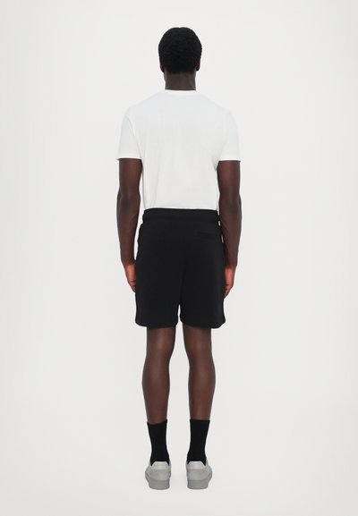 Schwarze Shorts mit einem elastischen Bund, getragen mit einem weißen T-Shirt und grauen Schuhen. Ansicht von hinten vor einem schlichten weißen Hintergrund.