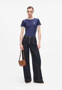 Marineblauwe geribde t-shirt met witte rand, gecombineerd met donkere wijde denim jeans met een ritssluiting aan de voorkant en contrasterende stiksels, aangevuld met een kleine bruine handtas.