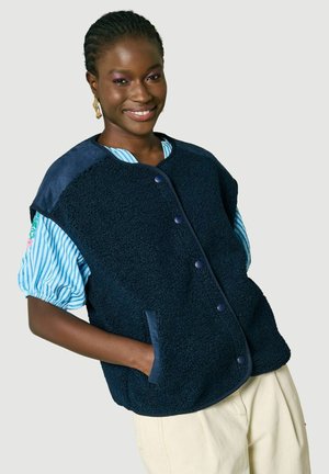 Donna sorridente con un gilet in pile blu navy sopra una camicia a maniche corte a righe blu e pantaloni color crema, in piedi davanti a uno sfondo neutro.