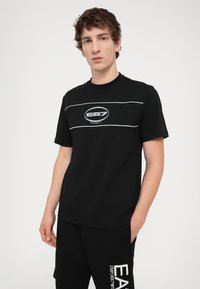 Black cotton t-shirt with a crew neck, featuring a white horizontal stripe and an oval EA7 logo on the chest. Short sleeves, relaxed fit.